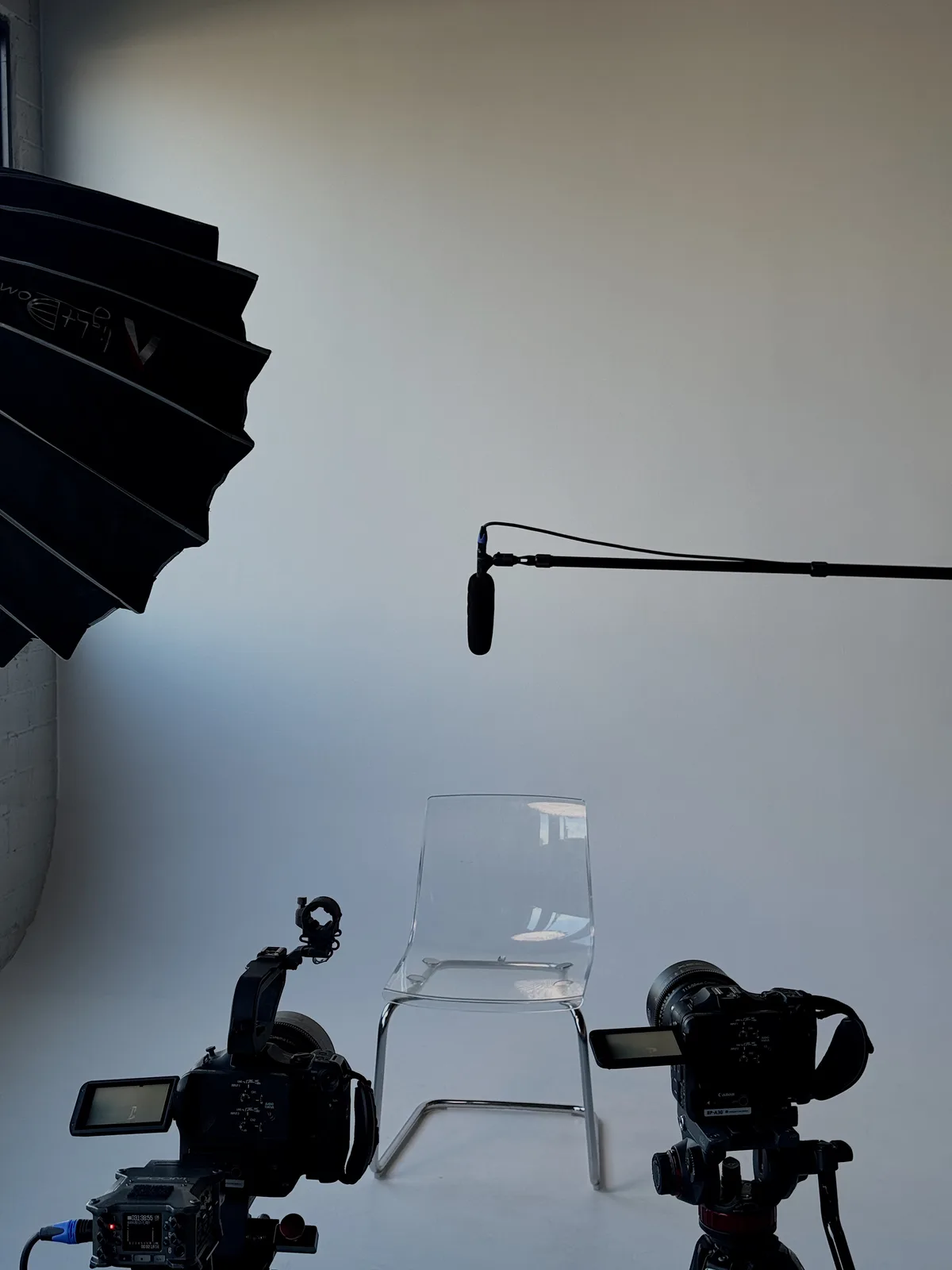 A clear acrylic chair set between two cameras and a boom microphone in front of a pale wall.