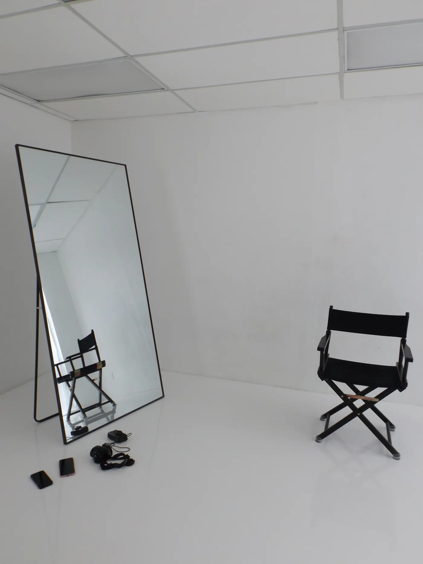 A full length mirror leaning in the corner of a white studio room, with a black director's chair across from it.